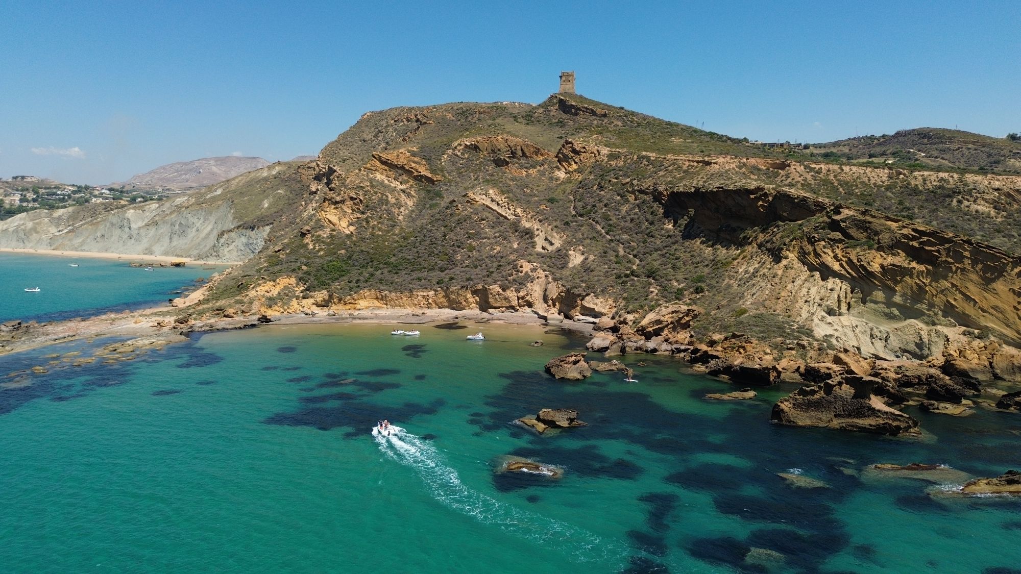 Una cala dall'alto, con le barche, gli scogli e una torre sul promontorio